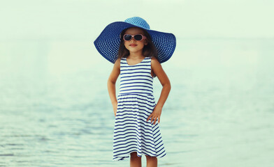 Summer portrait beautiful little girl child wearing a striped dress and straw hat on a beach on a sea background
