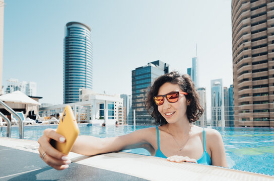 Cheerful woman travel blogger takes a selfie photo on her smartphone while relaxing in the pool on the roof of a skyscraper in Dubai
