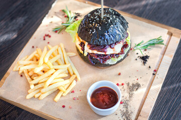 Black burger with French fries and ketchup on a wooden board in a restaurant. Restaurant menu