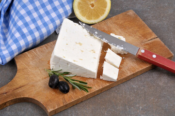 Morceau de feta sur une planche à découper avec un couteau, des olives noires, du citron et du romarin en gros plan