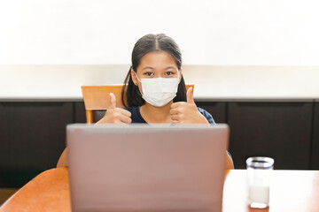 Little asian girl in face mask with thumb up study at home with laptop.