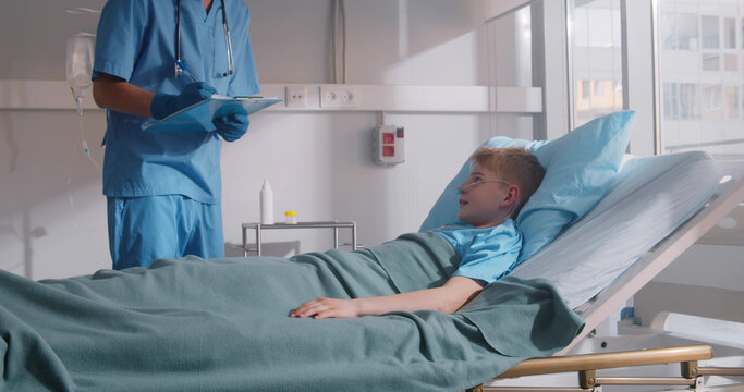 Cropped Shot Of Doctor Writing Notes Standing Near Hospital Bed Of Sick Boy Patient