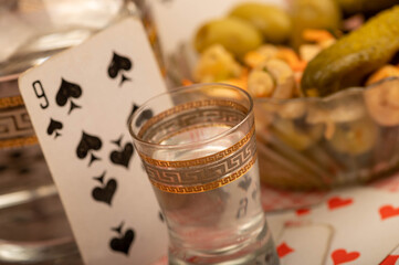 Playing cards, a glass of vodka, a decanter of vodka and a glass dish with pickles and mushrooms on the table.