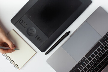 Top view photo of a female hand writing ona a notepad. A graphic design tablet, stylus and a laptop on a white background.