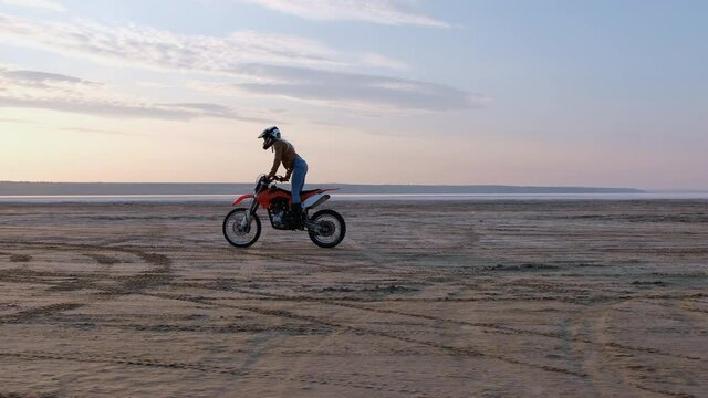 Sportive brave young woman does stunts standing on red motorbike riding on sand beach against boundless sea at sunset on horizon