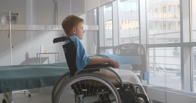Side view of pensive kid sitting on wheelchair looking out of window in hospital ward