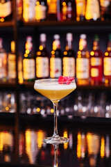 Elegant female yellow cocktail with white foam, decorated with pink flower petal on dark table against the background of blurred bar with expensive alcohol. Close-up. Space for inscriptions