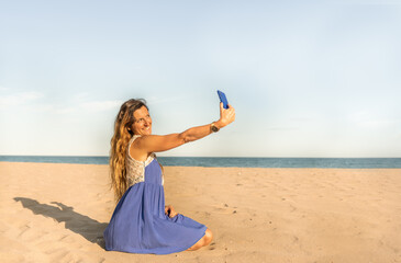 Fototapeta na wymiar Fashion woman taking a selfie with her mobile phone sitting on sandy beach at sunset.Summer lifestyle concept, female having fun using smartphone outdoor.