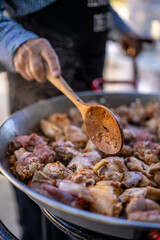 Hand of an old man chef cooking rabbit and chicken wings, pork ribs, in a paella, in outdoors and natural light, wearing a black apron, using a wooden big spoon