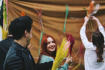 Obraz na płótnie Canvas young woman with red hair smiling at friend making arrangement with bouquet of flowers with brown background