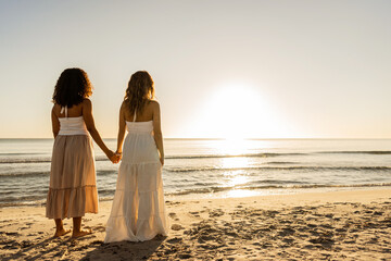 Couple of multiracial women in love view from back holding hands on the beach by the oceanic sea in...
