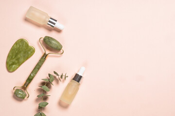 Jade roller and gua sha stone massager with serum bottles at pastel background. Anti-aging therapy. Top view with copy space.
