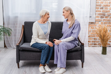 Obraz premium young social worker and senior woman smiling while talking on sofa at home