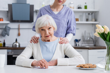 young nurse touching shoulders of elderly woman looking at camera near jigsaw puzzle