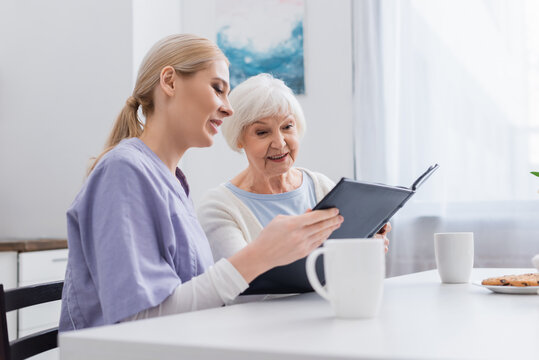 smiling nurse with happy elderly woman looking at photo album near cups of tea