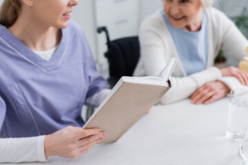 Obraz premium cropped view of young nurse reading book to elderly woman, blurred background