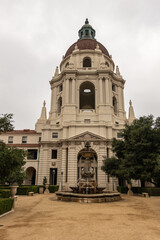 Fototapeta na wymiar Pasadena, CA, USA - May 11, 2021: Beige stone main tower with dome and fountain in front in sandy courtyard and green foliage garden of historic City Hall under silver sky. 