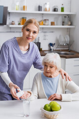 young nurse looking at camera while giving pills to aged woman in kitchen