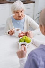Fototapeta premium senior woman talking to social worker near fresh apples and cups of tea, blurred foreground