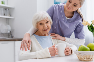 happy aged woman smiling at camera while young nurse hugging her shoulders in kitchen
