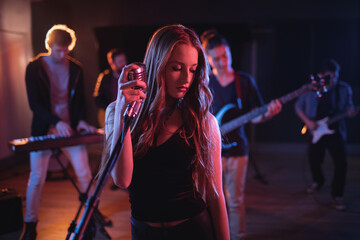 Caucasian female singer holding retro microphone at concert in music venue, music band behind her