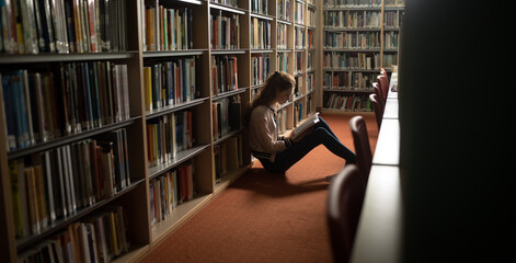 Caucasian female student with long hair leaning on bookshelf, reading book in library