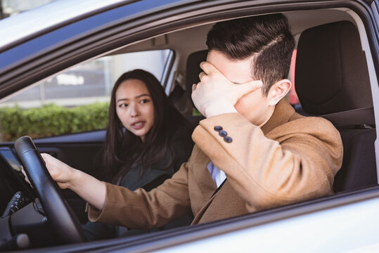 Diverse couple in car, man behind steering wheel covering his face woman looking at him