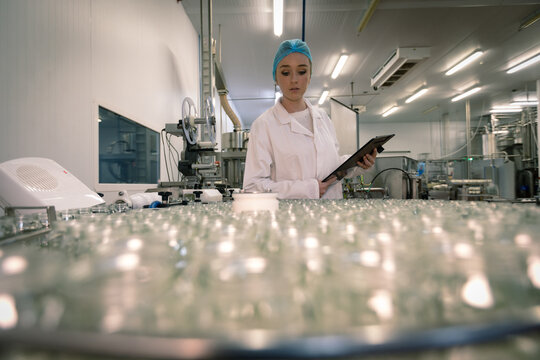 Caucasian woman wearing lab coat and cap holding clipboard inspecting glassware products in factory