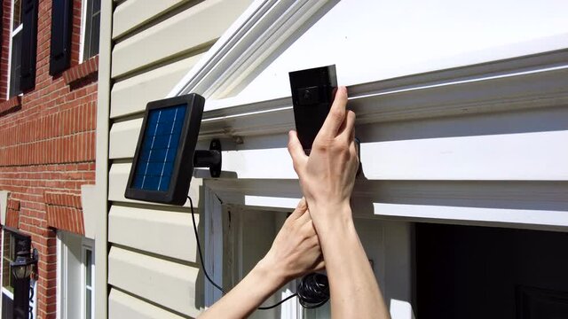 A caucasian woman is installing a wireless security surveilance camera with motion sensor on top of the front door frame. The camera is linked to a solar panel for environmental friendly charging. 