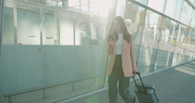 Busy young woman in formal clothes and eyeglasses walking with luggage and smartphone near airport. Business lady having working trip abroad. 