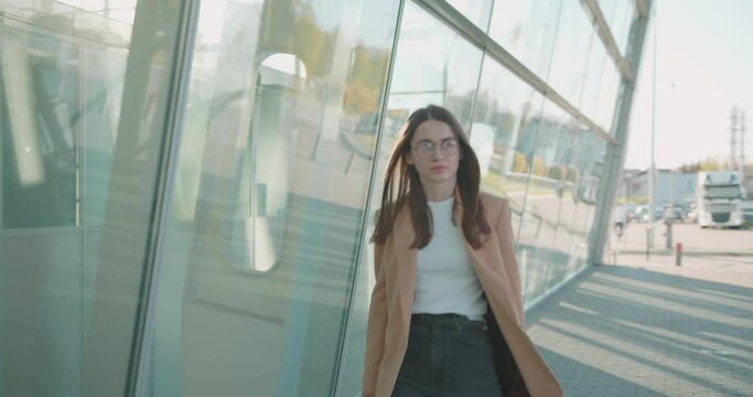 Confident business woman in eyeglasses and formal wear walking with suitcase outdoors. Modern glass airport on background. Business trip concept.