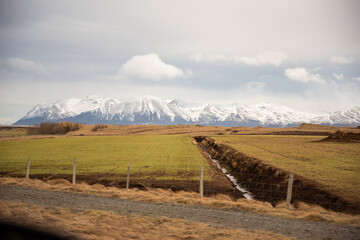 Iceland terrain of beautiful green and orange colors with waterway creating lines leading into a blue snow capped mountain range with puffy clouds dramatic heavenly farmland fencing