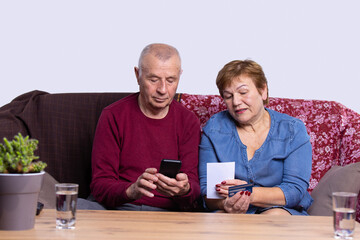 Serious elderly married couple paying for communal services through internet on mobile phone. Husband is sitting on sofa typing on device. Wife is dictating the credit card number