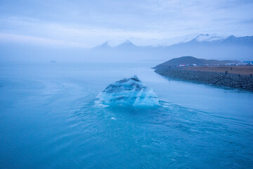 Icelandic glacier floating from global warming effects. Foggy atmosphere with snowcapped mountain range in the distance. Iceberg has layers of volcanic ash. © Jessica