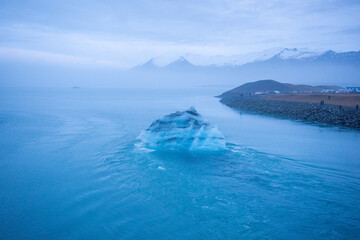 Icelandic glacier floating from global warming effects. Foggy atmosphere with snowcapped mountain range in the distance. Iceberg has layers of volcanic ash.