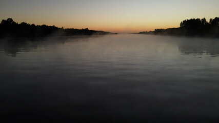 Foggy tranquil amazon river during colorful sunset in background. Silhouette of trees and plants on shore.
