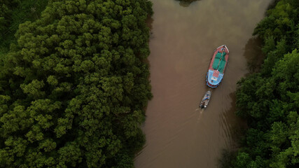 Top down shot of ship carrying small boat on amazon river surrounded by green rainforest trees during sunset.