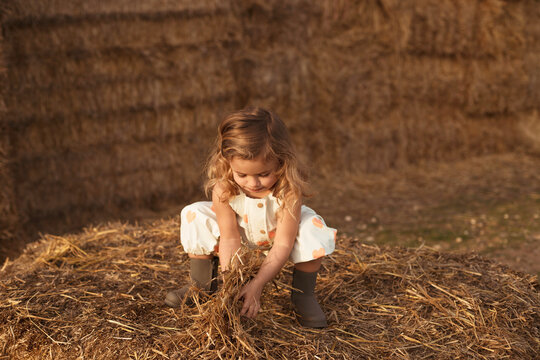 Side view of adorable child in overalls playing with hay near straw bales in countryside