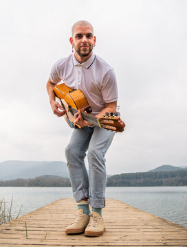 Full body of bearded guy in casual clothes standing with guitar on wooden pier near grass and river with mountains on background under cloudy gray sky in daytime