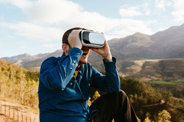 Curious male traveler sitting on hill and experiencing virtual reality in goggles in mountains on sunny day