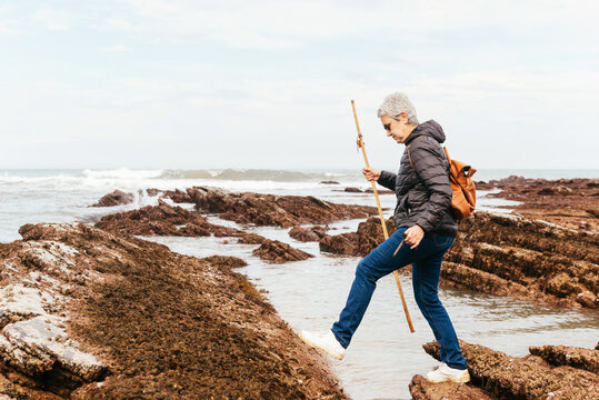 Side view of elderly female backpacker with trekking pole strolling on boulders against stormy ocean under cloudy sky