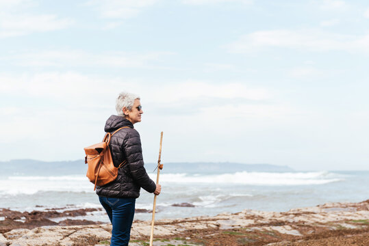 Side view of elderly female backpacker with trekking pole strolling on boulders against stormy ocean under cloudy sky