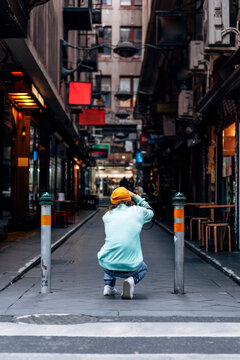 Back view of unrecognizable man in casual outfit squatting on pavement while taking photo of urban buildings on camera