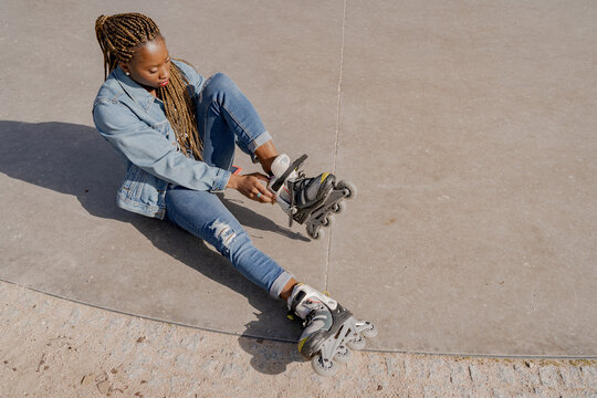 From above of African American female in denim outfit putting on rollerblades on sunny day in skate park