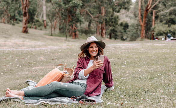 Cheerful female in hat sitting on blanket on meadow in forest and browsing mobile phone while enjoying picnic in Australia
