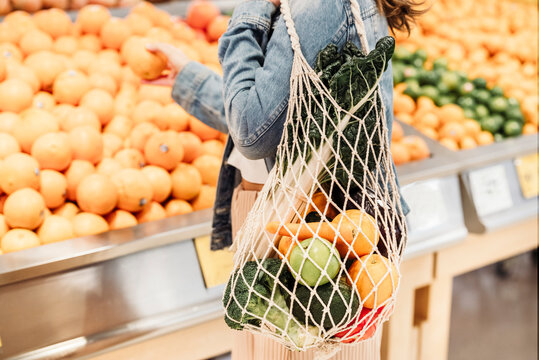 Side view of crop unrecognizable female buyer standing in supermarket with eco friendly mesh bag full of ripe fruits and vegetables