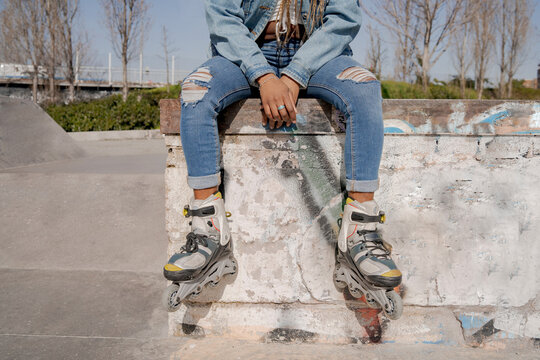 Crop black female with braided hairstyle and in rollerblades sitting on ramp in skate park and looking away
