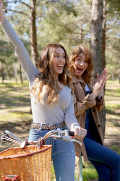 Happy young girlfriends smiling and looking at camera on bicycle in sunny park in summer