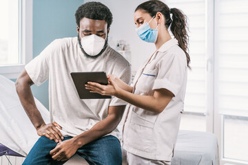Young female doctor in medical uniform and stethoscope wearing face mask speaking and showing result on tablet to African American man patient during appointment in clinic