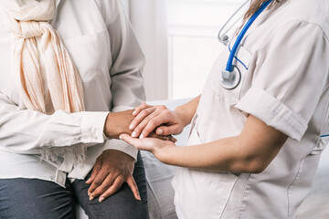 Cropped unrecognizable female doctor in medical uniform and stethoscope wearing face mask speaking and holding hands of African American mature woman patient during appointment in clinic